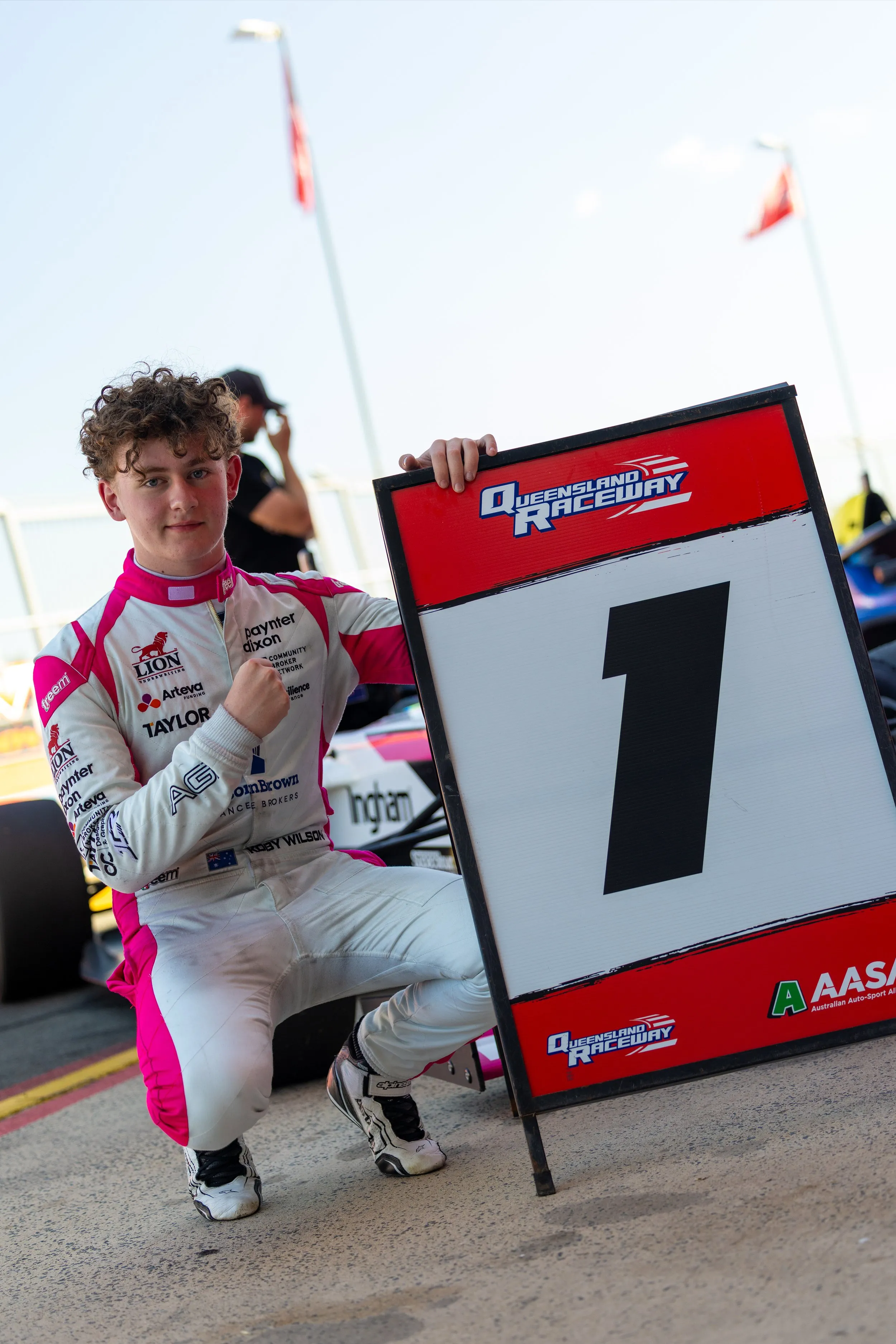 Koby crouching beside the number 1 board at Queensland Raceway, fist pressed to his chest