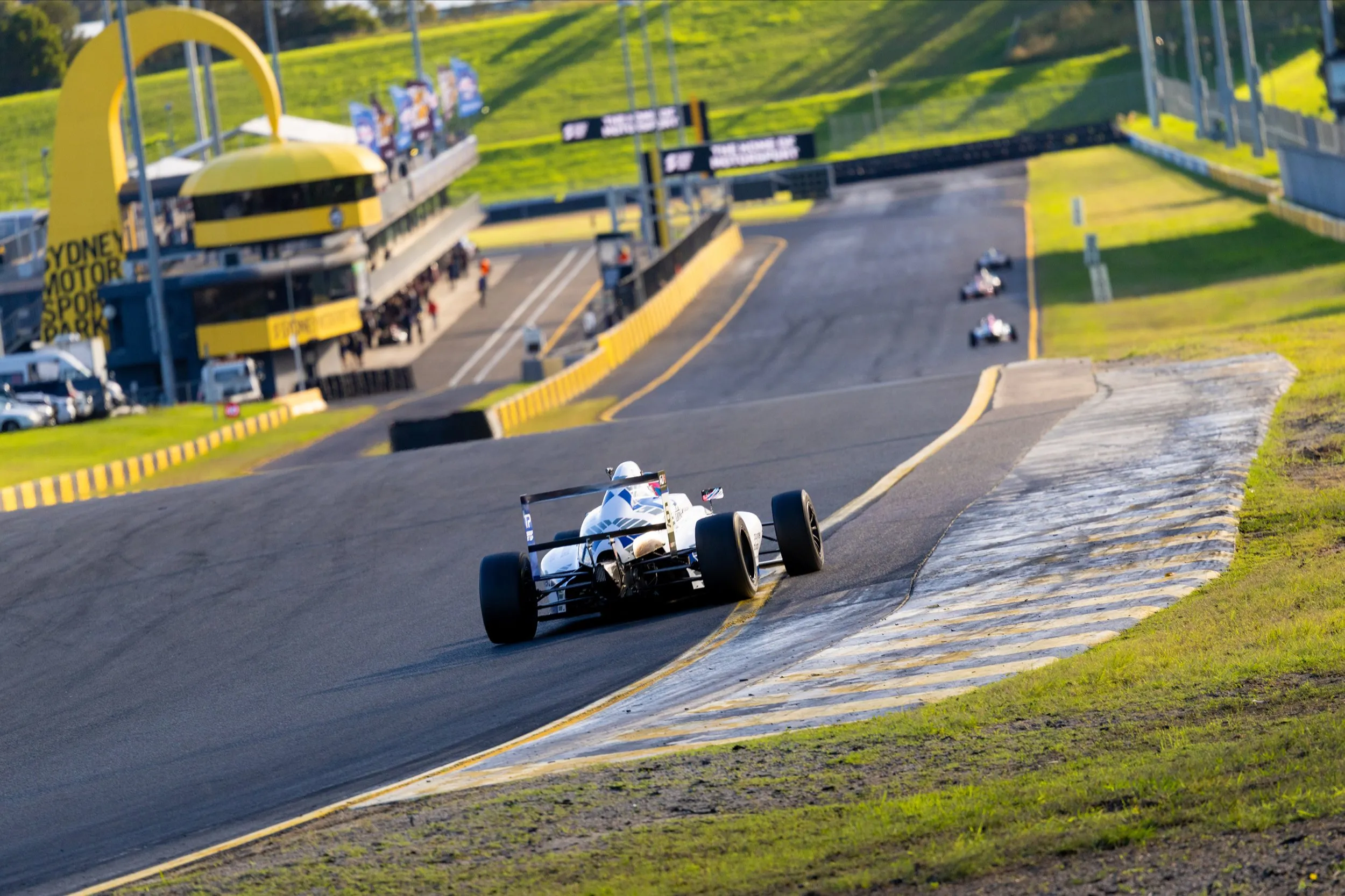 Koby's car climbing the hill at Sydney Motorsport Park in late-afternoon light, with the circuit's yellow arch behind