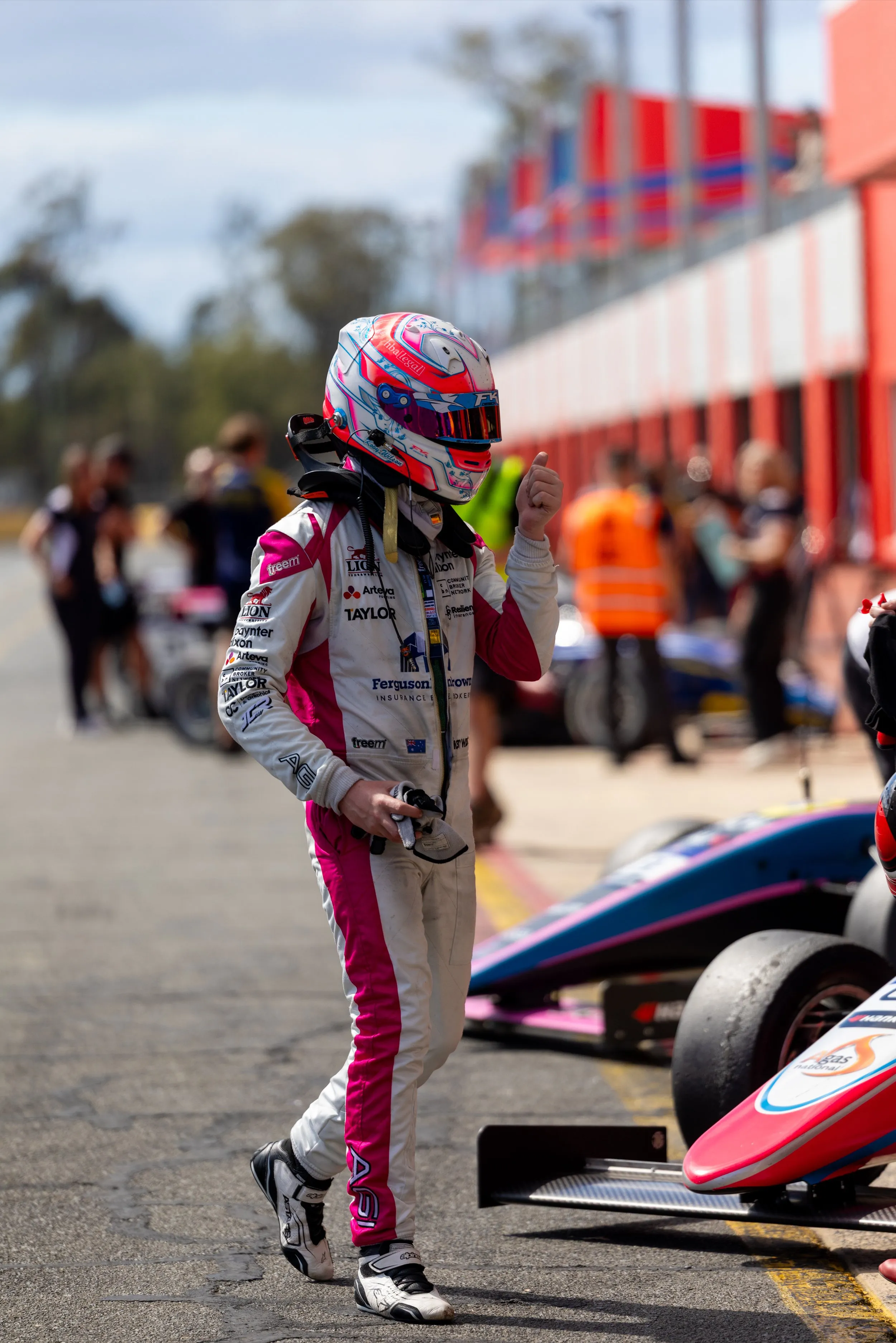 Koby walking along the pit lane in his race suit and helmet, past parked cars and crew